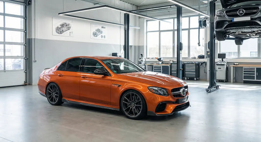 Fotorealistischer E63 W213 in orange, leicht seitlich von vorne fotografiert, steht in einer sauberen, modern beleuchteten Werkstatt mit Hebebühne im Hintergrund. 